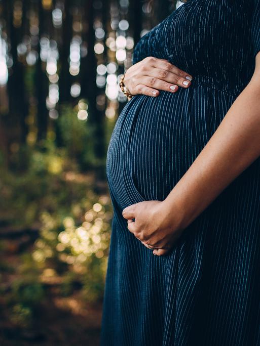 Eine schwangere Frau hält ihren Bauch. Sie steht in einem Wald. Das Licht bricht durch die Blätter. Ihr Gesicht ist nicht zu sehen (Symbolbild).