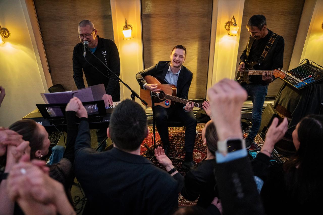 Ein lächelnder Mann mit Gitarre wird auf der Bühne von zwei Musikern begleitet.