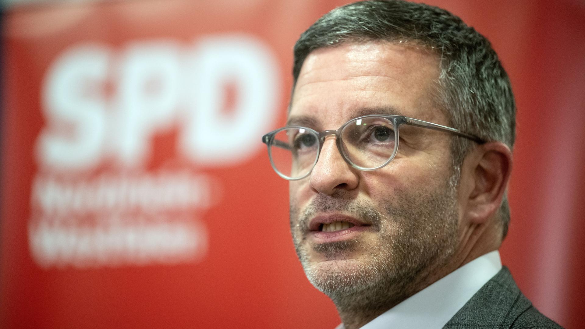 Herter im dunklen karierten Jackett und mit Brille im Vordergrund, im Hintergrund verschwommen das rot-weiße Logo der SPD.