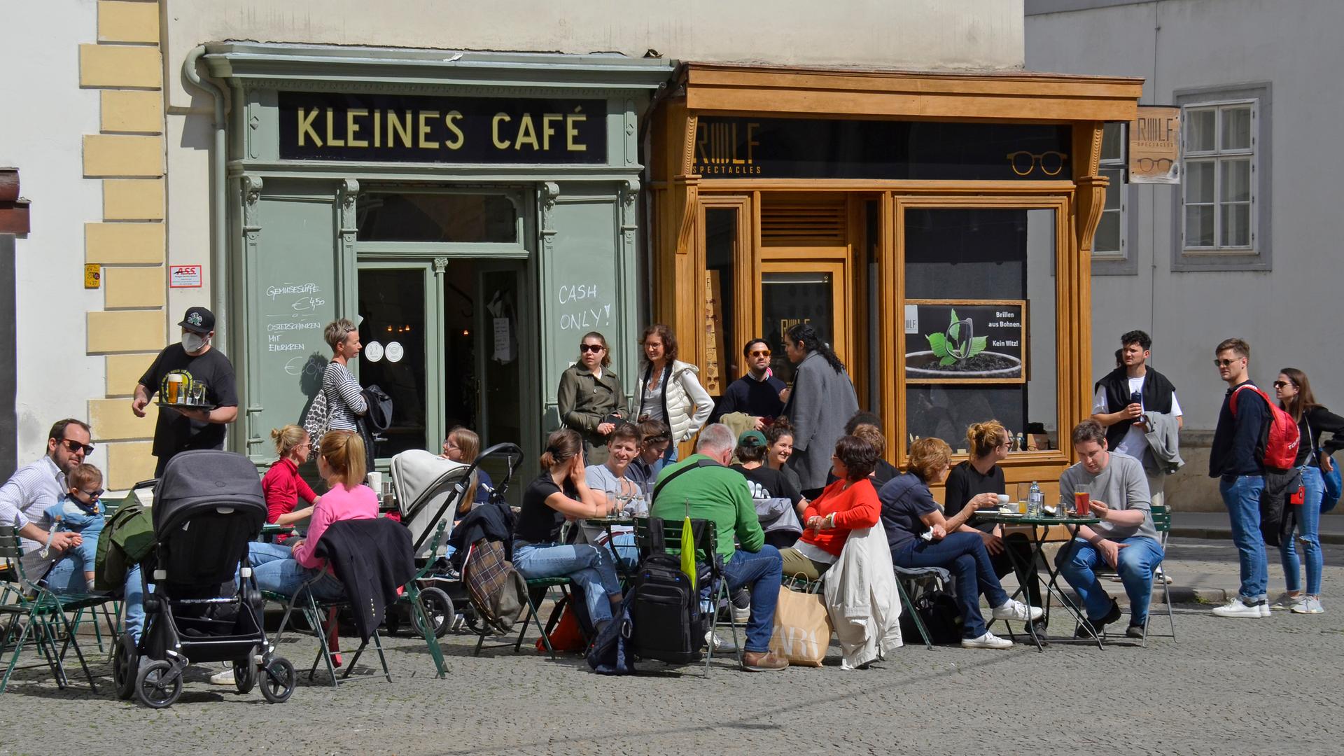 Das Kleine Café von außen, davor sitzen mit salbeigrüner Fassade von außen, Gäste sitzen davor an kleinen Tischen in der Sonne. 