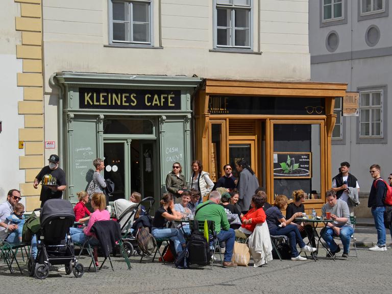 Das Kleine Café von außen, davor sitzen mit salbeigrüner Fassade von außen, Gäste sitzen davor an kleinen Tischen in der Sonne. 