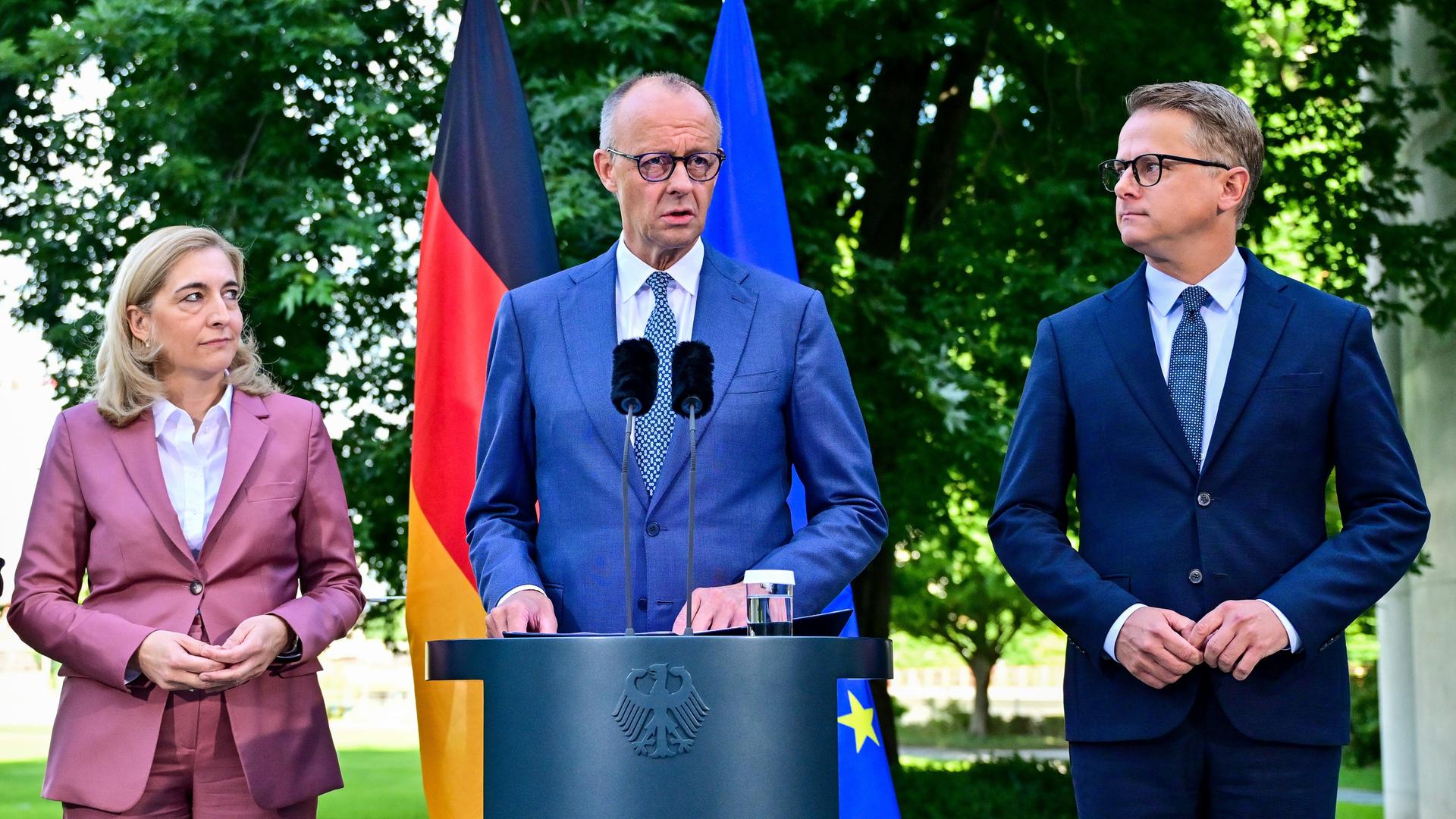 Das Bild zeigt Bundes-Kanzler Friedrich Merz, die Chefin von dem Kanzler-Amt Nina Warken und den Gesundheits-Minister Carsten Linnemann. Die Politiker stehen zusammen und geben eine Presse-Konferenz. Hinter den Politikern stehen zwei Fahnen. Das Bild zeigt Bundes-Kanzler Friedrich Merz, die Chefin von dem Kanzler-Amt Nina Warken und den Gesundheits-Minister Carsten Linnemann. Die Politiker stehen zusammen und geben eine Presse-Konferenz. Hinter den Politikern stehen zwei Fahnen.