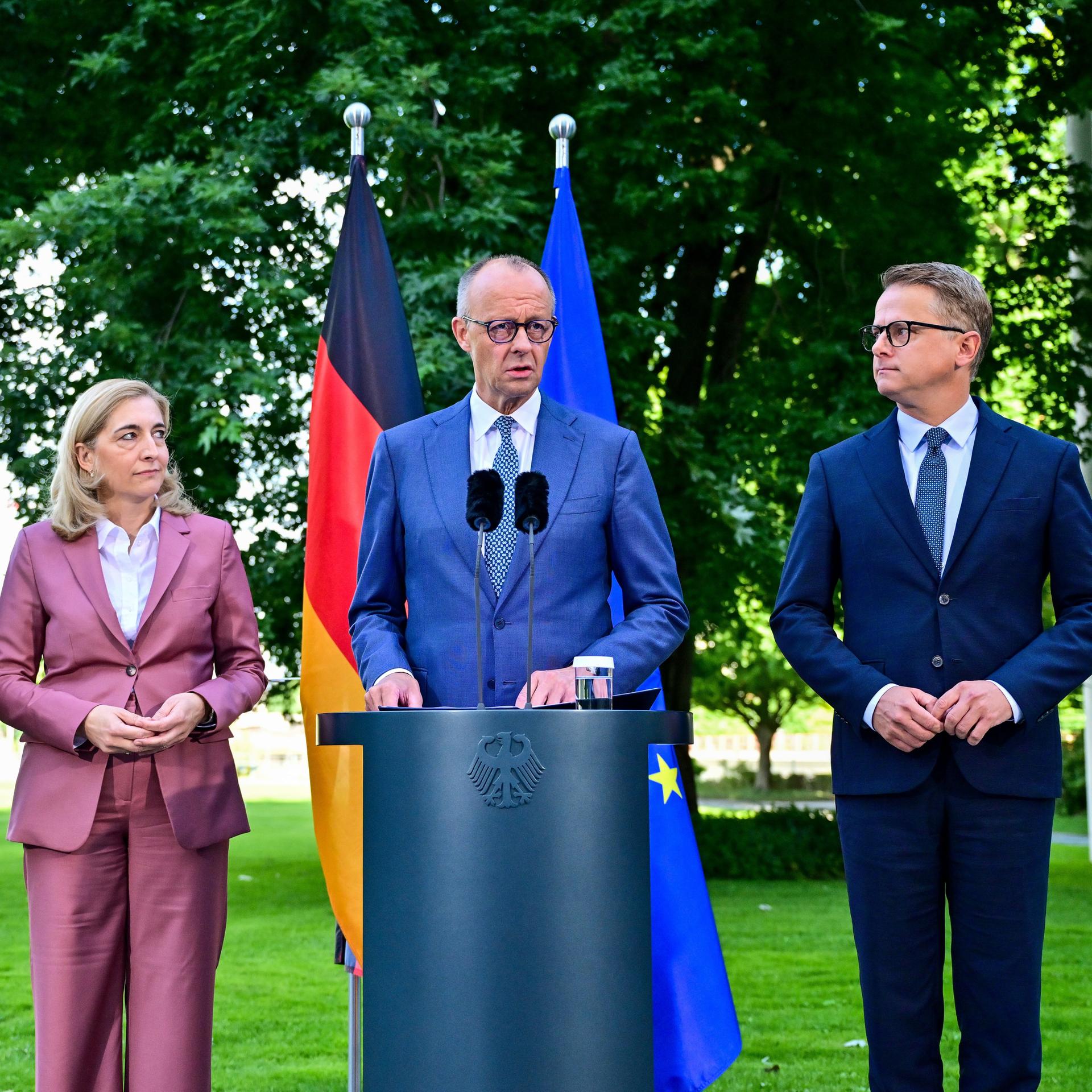 Das Bild zeigt Bundes-Kanzler Friedrich Merz, die Chefin von dem Kanzler-Amt Nina Warken und den Gesundheits-Minister Carsten Linnemann. Die Politiker stehen zusammen und geben eine Presse-Konferenz. Hinter den Politikern stehen zwei Fahnen.