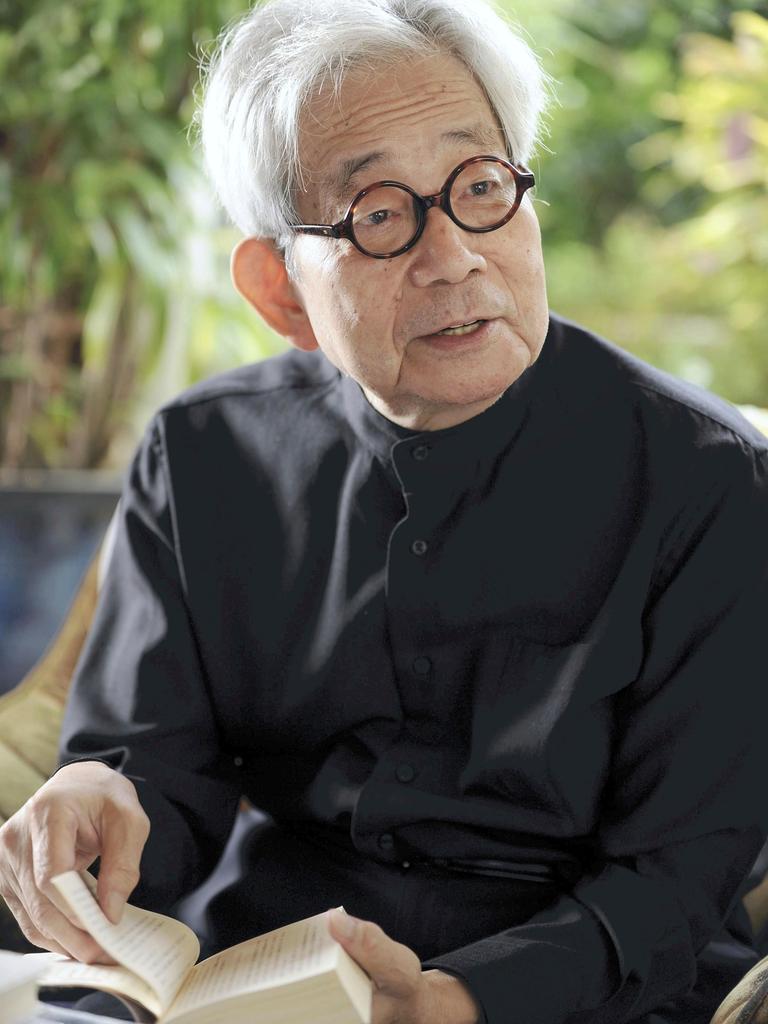 Kenzaburo Oe während eines Interviews. Er trägt ein schwarzes Hemd mit Mandarinkragen, eine runde Hornbrille und hält ein Buch aufgeschlagen in der Hand.