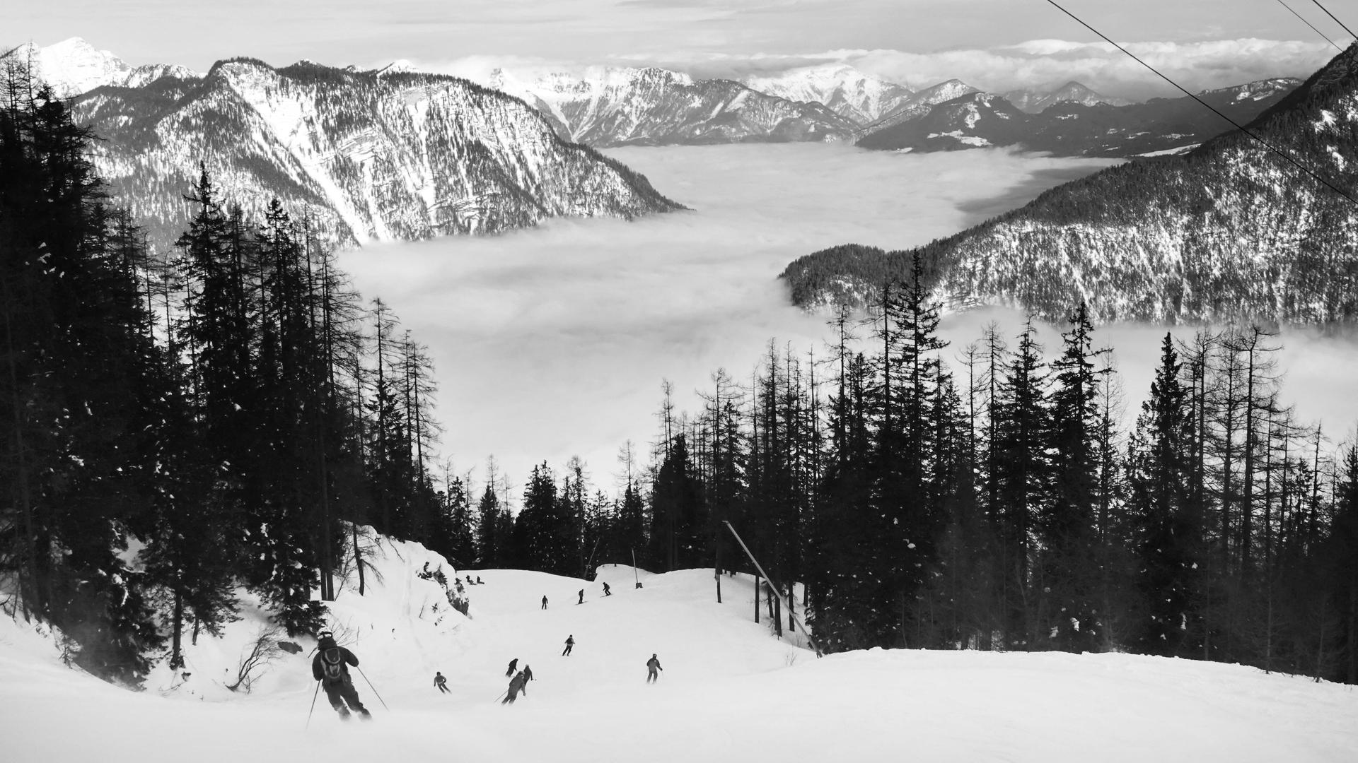 Schwarz-weiß Foto: Menschen fahren Ski am Krippenstein. Blick auf die Piste und das Panorama, wolkenverhangende Berge.