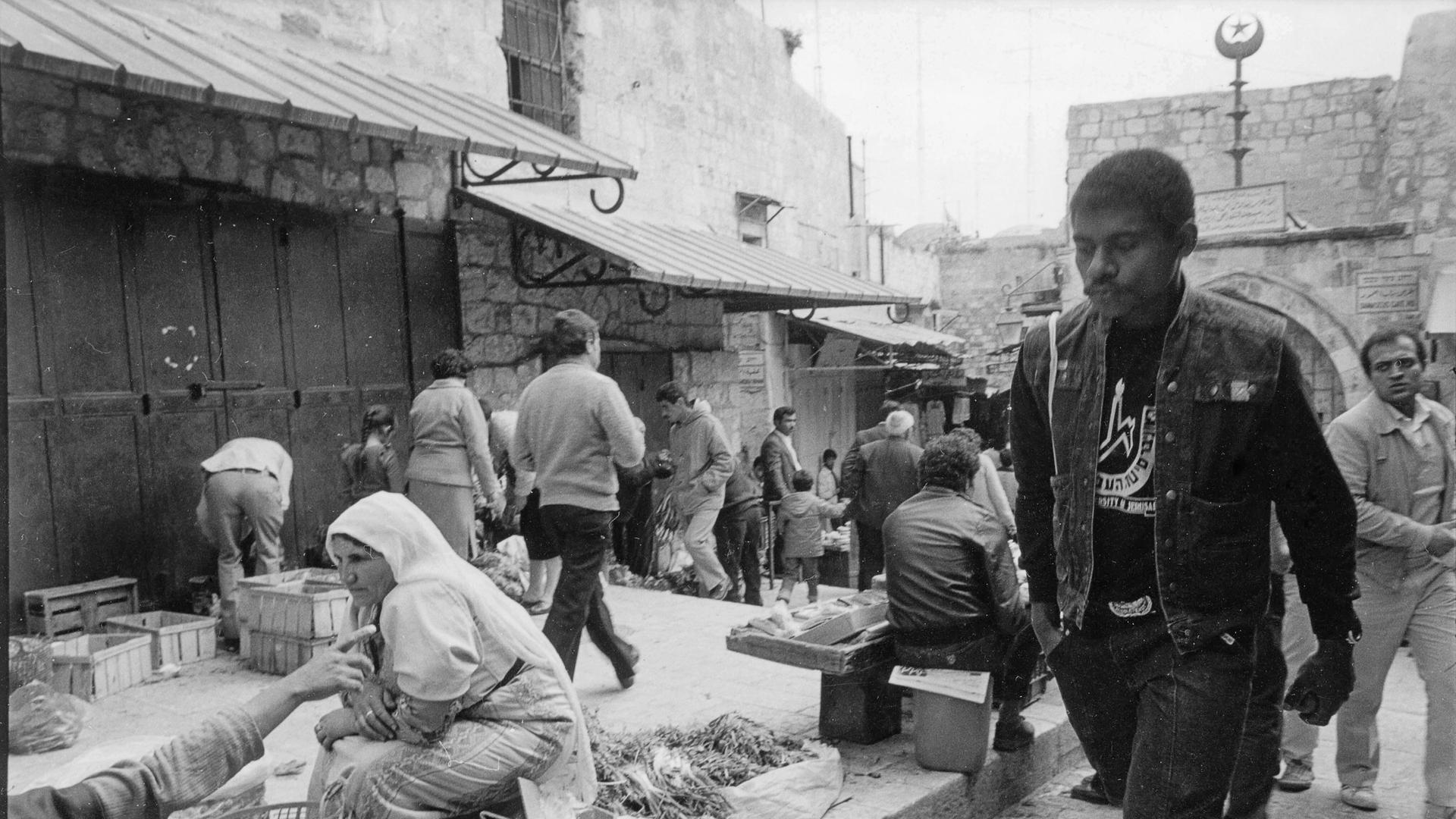 Historische Schwarz-Weiss Fotografie mit einer Straßenszene aus Jerusalem. Ein junger schwarzer Mann in Jeansweste geht entlang einer Marktraße.
