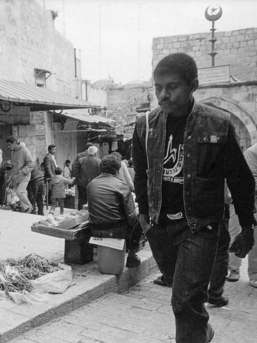 Historische Schwarz-Weiss Fotografie mit einer Straßenszene aus Jerusalem. Ein junger schwarzer Mann in Jeansweste geht entlang einer Marktraße.
