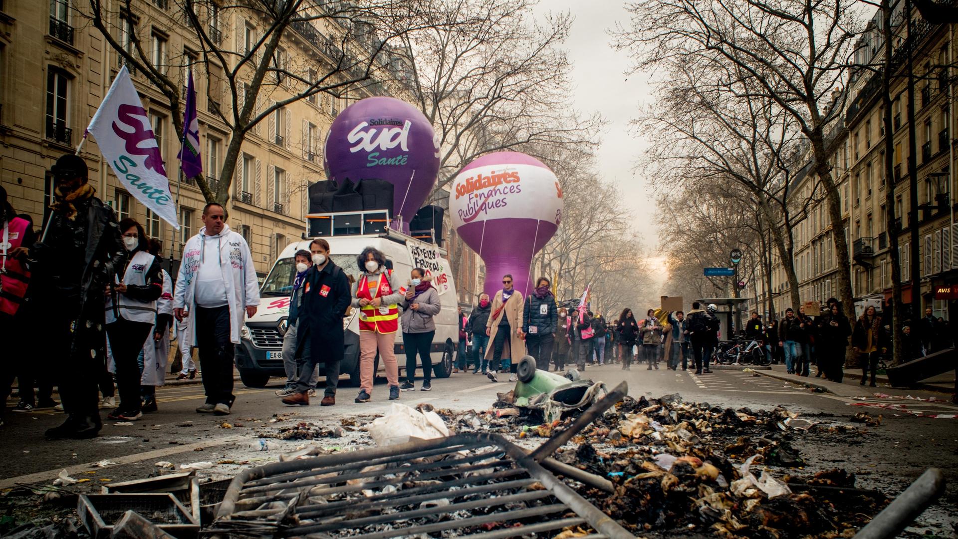 Ein Demonstrationszug in Paris, im Vordergrund des Bildes reste eines Brandes und ein liegendes Absperrgitter.