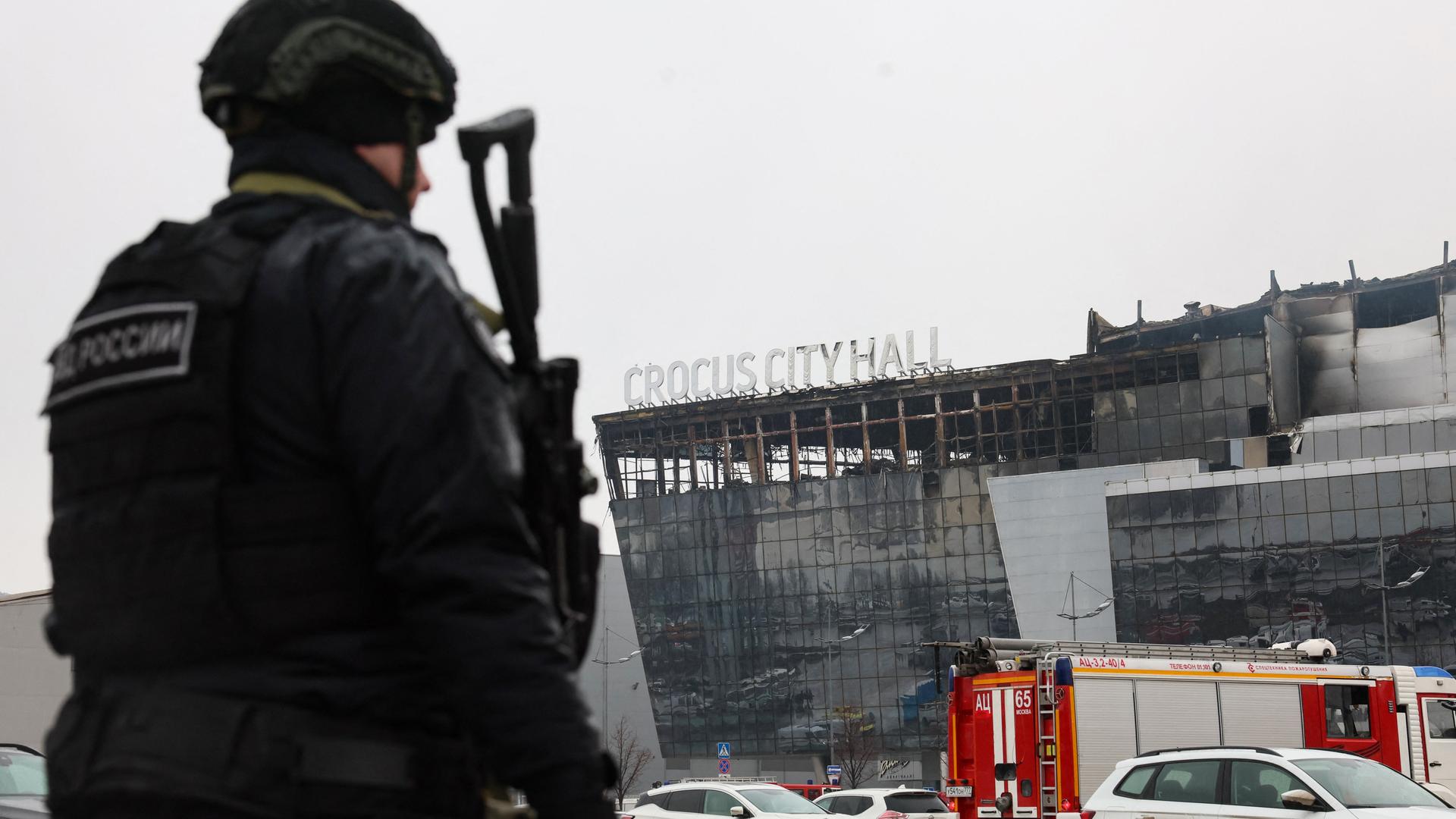 Ein Polizeibeamter patrouilliert am 23. März 2024 am Ort des Schusswaffenangriffs in der Konzerthalle „Crocus City Hall“ in Krasnogorsk bei Moskau. Im Vordergruns ist der Polizist in voller Montur zu sehen, im Hintergrund die abgebrannte Stadthalle.