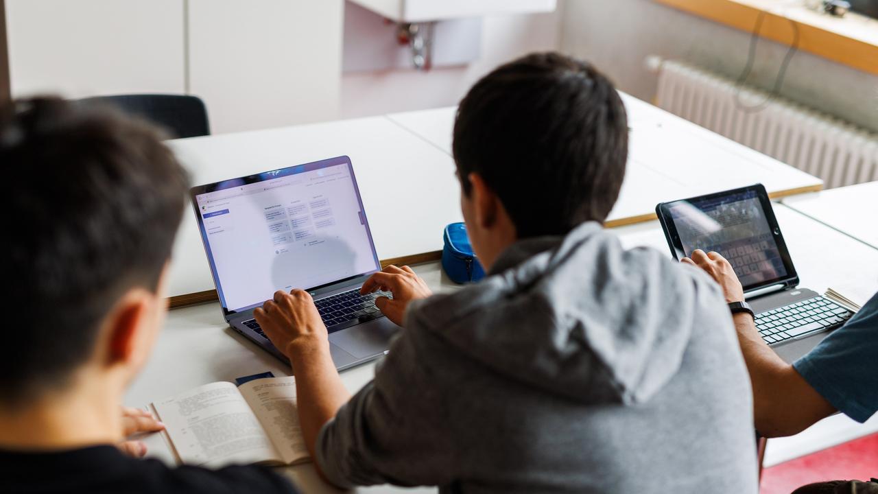 Drei Schüler von hinten an einer Schulbank; zwei von ihnen schauen in ein Tablet.