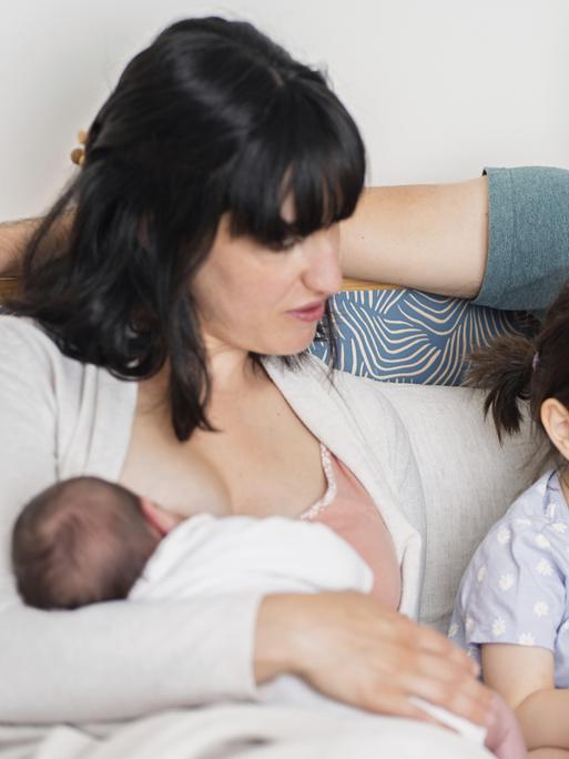 Eine junge Familie sitzt zuhause auf dem Sofa. Die Mutter hält ein neugeborenes Baby in der Hand, während der Vater und die große Schwester lesen Eine junge Familie sitzt zuhause auf dem Sofa. Die Mutter hält ein neugeborenes Baby in der Hand, während der Vater und die große Schwester lesen