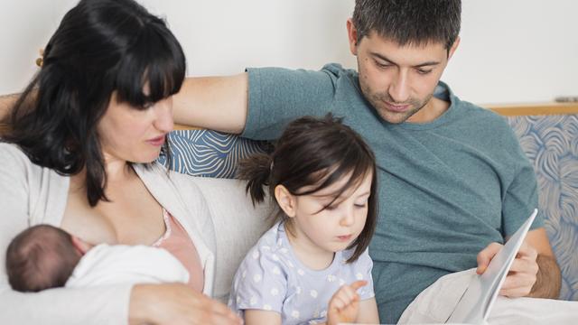 Eine junge Familie sitzt zuhause auf dem Sofa. Die Mutter hält ein neugeborenes Baby in der Hand, während der Vater und die große Schwester lesen