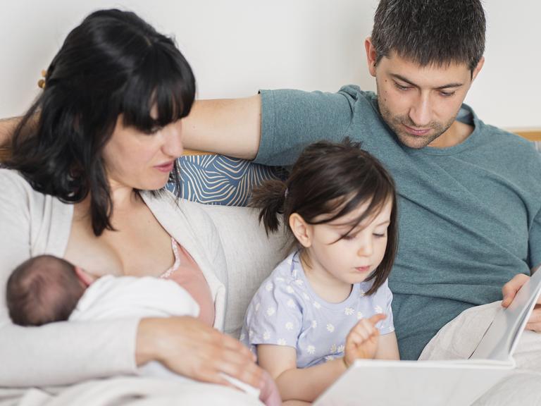Eine junge Familie sitzt zuhause auf dem Sofa. Die Mutter hält ein neugeborenes Baby in der Hand, während der Vater und die große Schwester lesen Eine junge Familie sitzt zuhause auf dem Sofa. Die Mutter hält ein neugeborenes Baby in der Hand, während der Vater und die große Schwester lesen