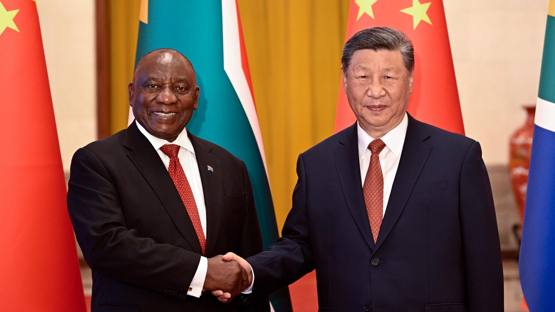 Xi Jinping und Cyril Ramaphosa geben sich die Hand und schauen lächelnd in die Kamera. Im Hintergrund sind die Flaggen Chinas und Südafrikas zu sehen. Xi Jinping und Cyril Ramaphosa geben sich die Hand und schauen lächelnd in die Kamera. Im Hintergrund sind die Flaggen Chinas und Südafrikas zu sehen.