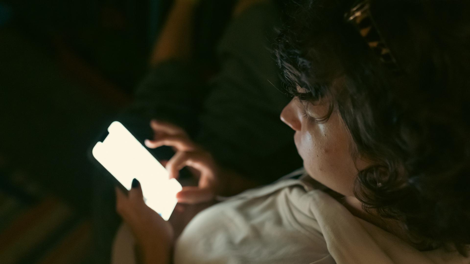 Ein Kind sitzt in dunkler Umgebung und schaut auf das Display seines Smartphones. Ein Kind sitzt in dunkler Umgebung und schaut auf das Display seines Smartphones.
