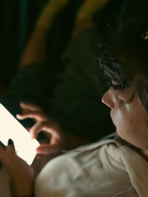 Ein Kind sitzt in dunkler Umgebung und schaut auf das Display seines Smartphones. Ein Kind sitzt in dunkler Umgebung und schaut auf das Display seines Smartphones.