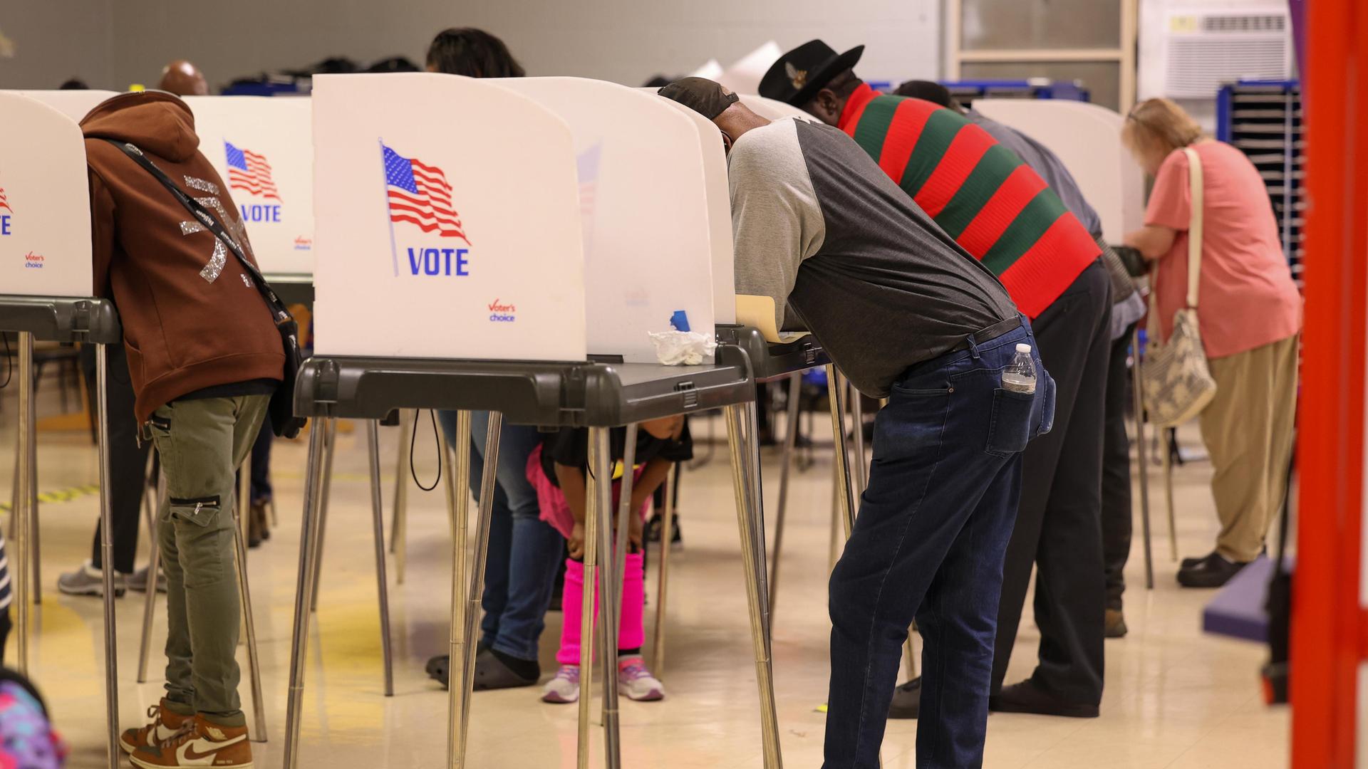 Wählerinnen und Wähler stehen an hohen abgeschirmten Tischen, die als Wahlkabinen fungieren. Auf der Seitenwand der Kabine ist die Flagge der USA zu sehen. Darunter der Schriftzug „Vote“. 