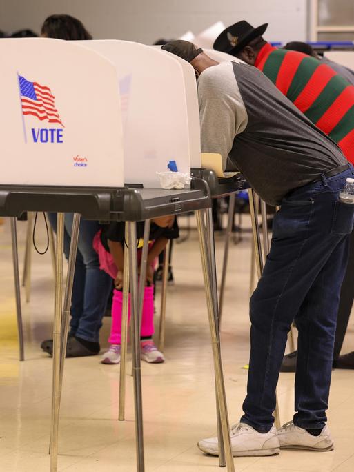 Wählerinnen und Wähler stehen an hohen abgeschirmten Tischen, die als Wahlkabinen fungieren. Auf der Seitenwand der Kabine ist die Flagge der USA zu sehen. Darunter der Schriftzug „Vote“. 