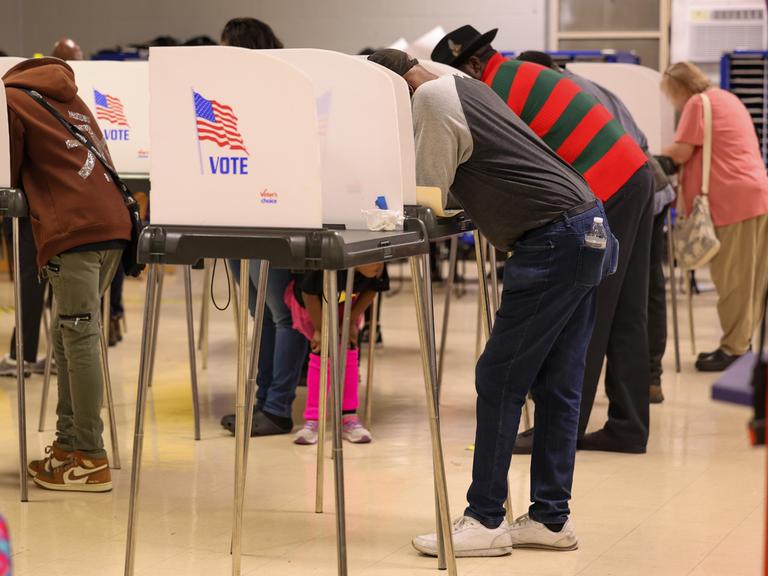 Wählerinnen und Wähler stehen an hohen abgeschirmten Tischen, die als Wahlkabinen fungieren. Auf der Seitenwand der Kabine ist die Flagge der USA zu sehen. Darunter der Schriftzug „Vote“. 