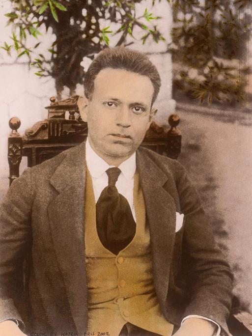 Eine historische, nachkolorierte Fotoaufnahme zeigt Kurt Tucholsky sitzend und ernst in die Kamera blickend.