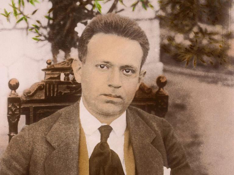 Eine historische, nachkolorierte Fotoaufnahme zeigt Kurt Tucholsky sitzend und ernst in die Kamera blickend.