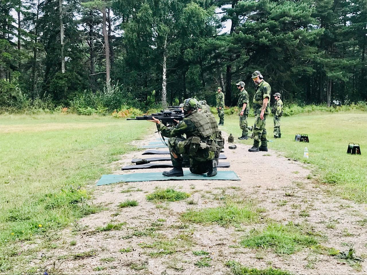Frauen und Männer knien in militärischer Tarnkleidung auf Matten auf einer Wiese, die Gewehre im Anschlag. Ihre Trainer stehen hinter ihnen.
