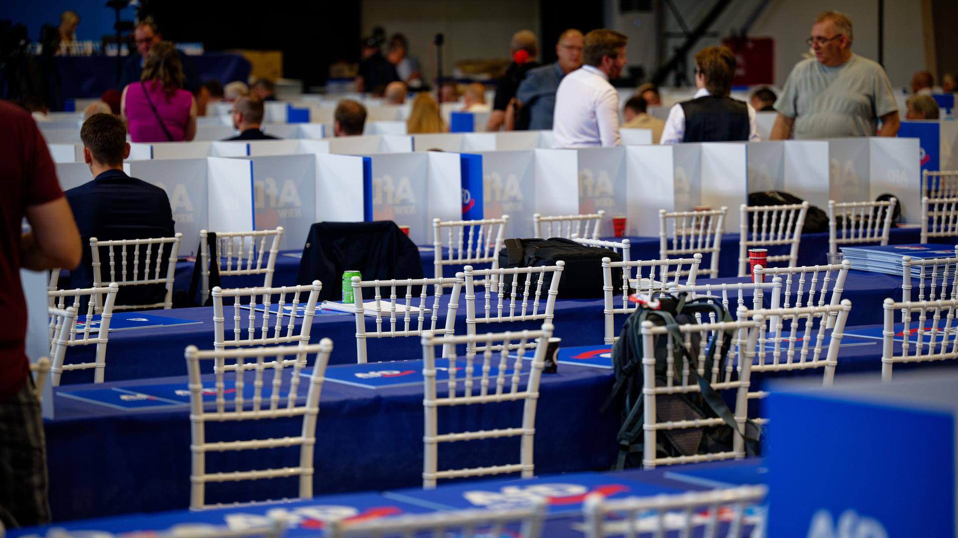 Das Bild zeigt unbesetzte Stühle in dem Saal des Parteitags. Im Hintergrund sind Delegierte der AfD zu sehen.