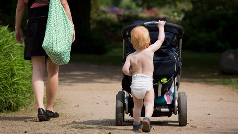 Ein Kleinkind mit Windel schiebt seinen Kinderwagen durch einen Park.