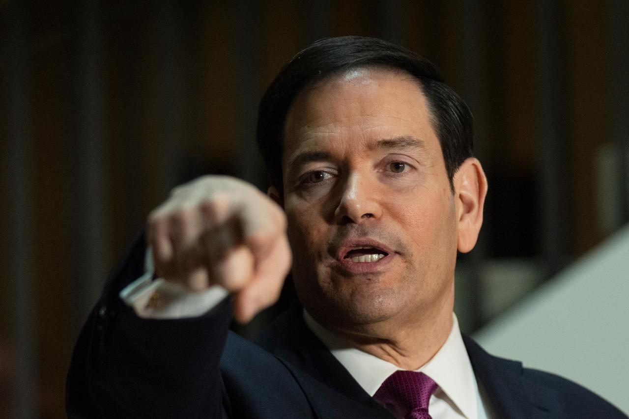 Das Bild zeigt den US-Außenminister Marco Rubio. Er ist ein Mann mittleren Alters mit dunklen Haaren und einem dunklen Anzug. Er gestikuliert mit der Hand. Das Bild zeigt den US-Außenminister Marco Rubio. Er ist ein Mann mittleren Alters mit dunklen Haaren und einem dunklen Anzug. Er gestikuliert mit der Hand.