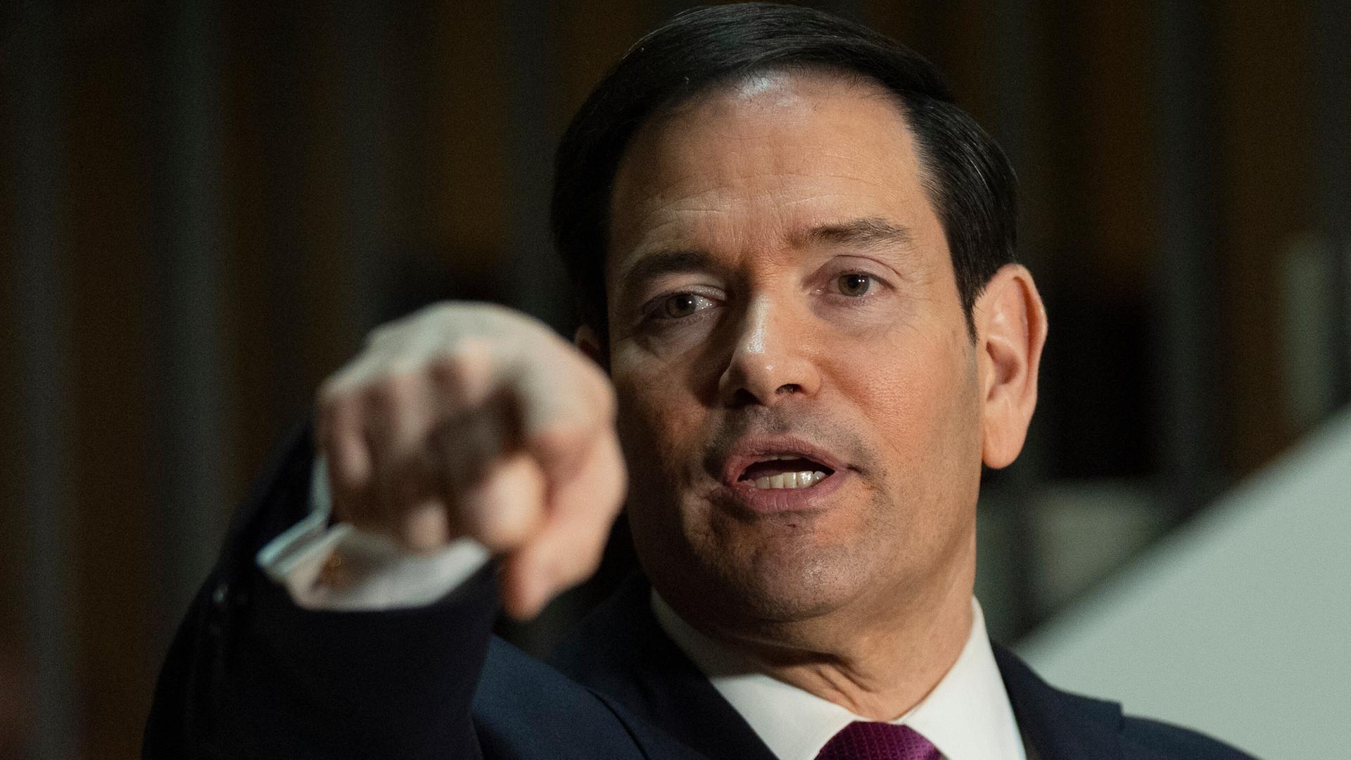 Das Bild zeigt den US-Außenminister Marco Rubio. Er ist ein Mann mittleren Alters mit dunklen Haaren und einem dunklen Anzug. Er gestikuliert mit der Hand.