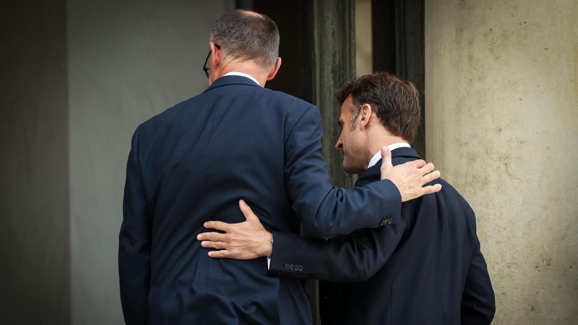 Rückenansicht des französischen Präsidenten Emmanuel Macron und des Bundeskanzlers Friedrich Merz. Beide haben sich freundschaftlich die Arme um die Schulter gelegt. 