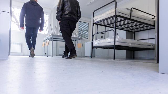 Blick aus niedriger Perspektive auf den weißen Binde des Raums mit mehreren Stockbetten. Zwei Männer, die man von hinten sieht, gehen gerade hinein. 