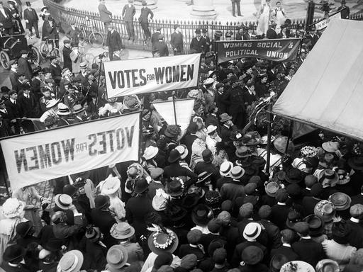 In London demonstrieren im Jahr 1910 Frauenrechtlerinnen vor der Queen's Hall. Sie tragen Banner, die das Wahlrecht für Frauen einfordern. 
