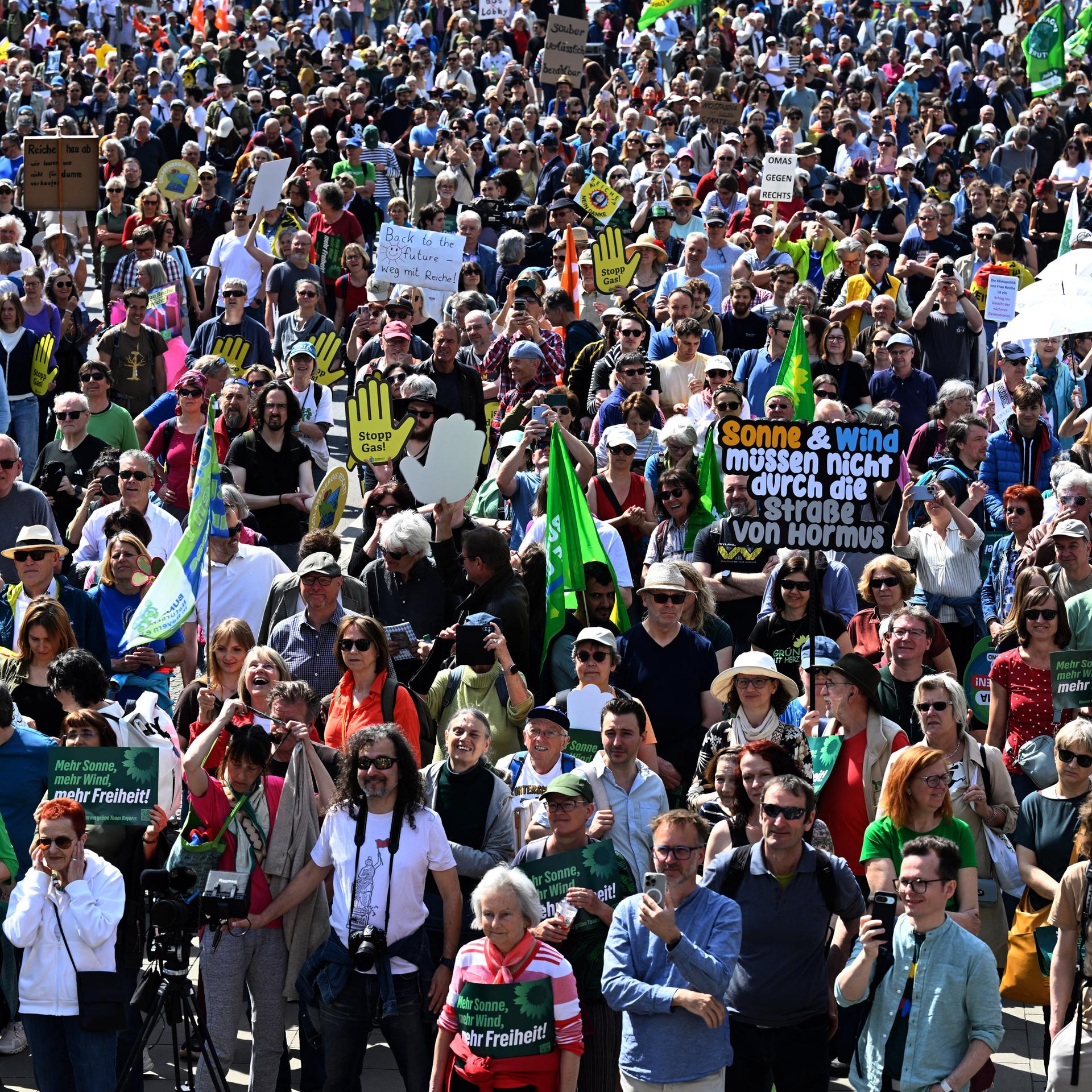 Teilnehmer und Teilnehmerinnen stehen in München am Siegestor. Sie halten Plakate - zum Beispiel mit der Aufschrift "Sonne und Wind müssen nicht durch die Straße von Hormus".