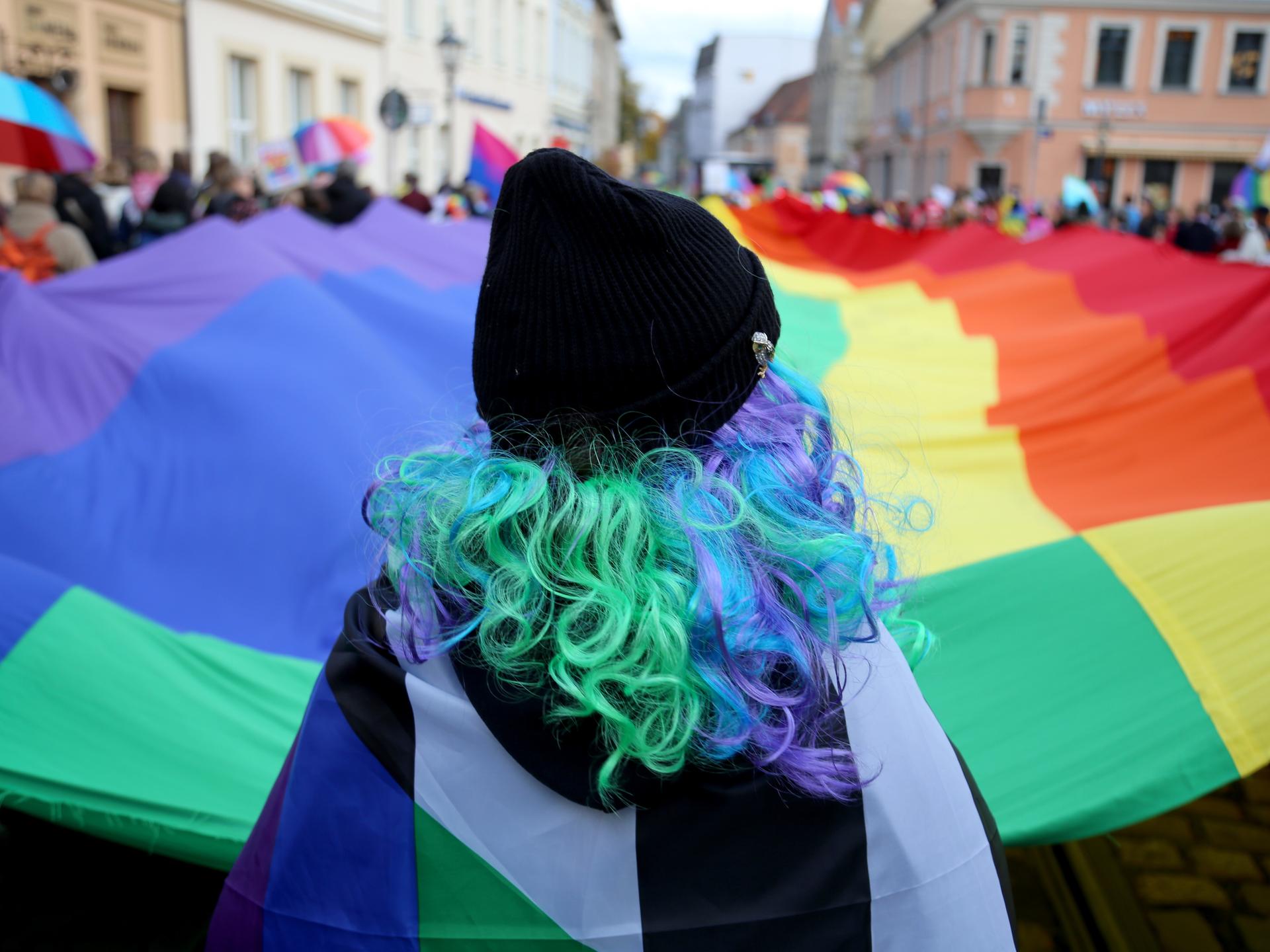 Teilnehmer schwenken eine überdimensionale Regenbogenflagge während des Christopher Street Day in Cottbus. Im Vordergrund ist eine Person mit grünen gelockten Haaren von hinten zu sehen.