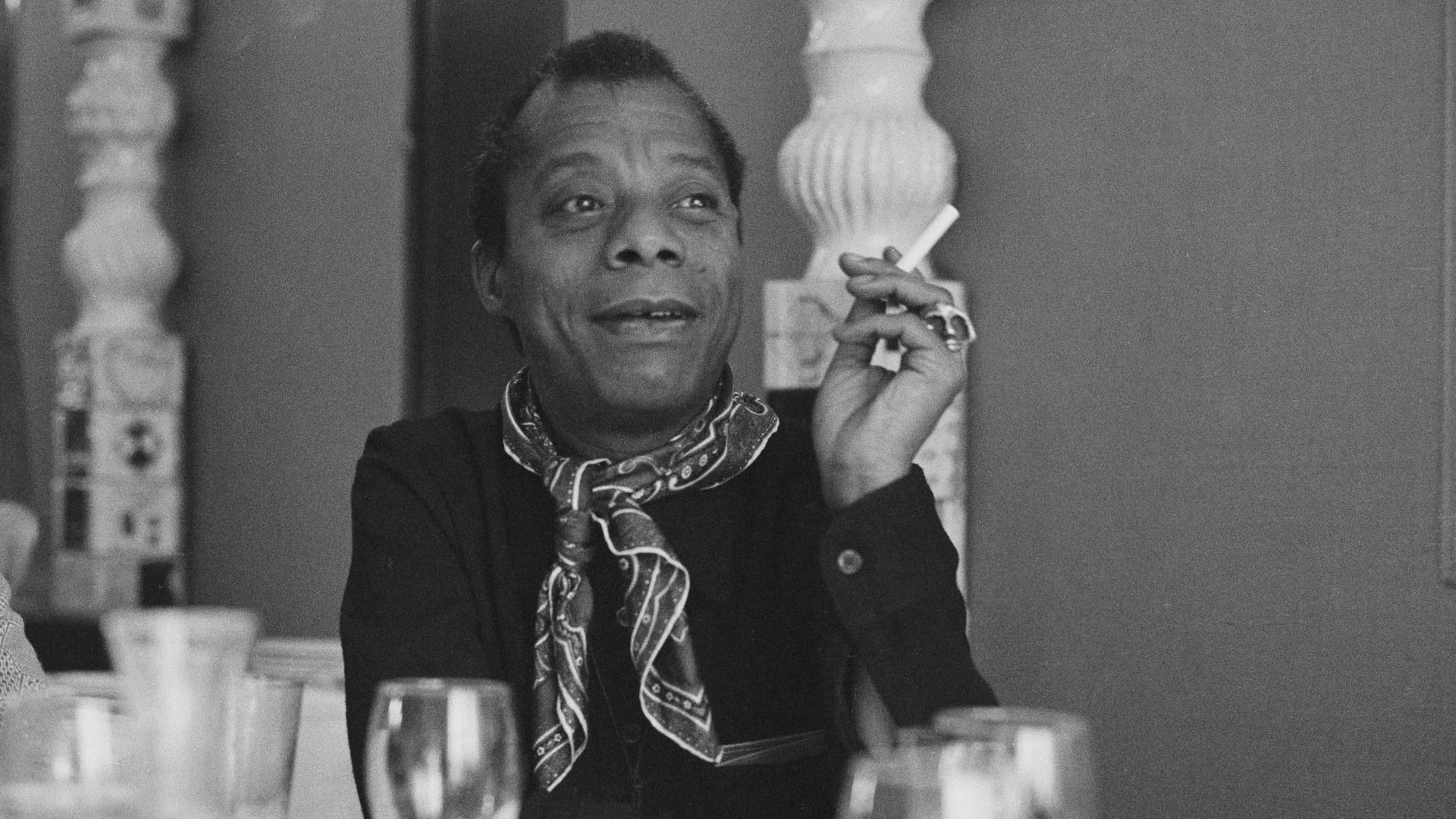 James Baldwin sitzt an einem gedeckten Tisch, raucht eine Zigarette und schaut zur Seite.