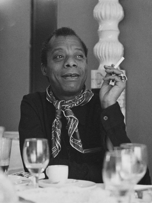 James Baldwin sitzt an einem gedeckten Tisch, raucht eine Zigarette und schaut zur Seite.