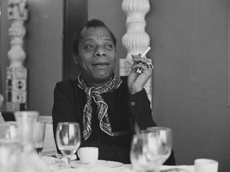 James Baldwin sitzt an einem gedeckten Tisch, raucht eine Zigarette und schaut zur Seite.