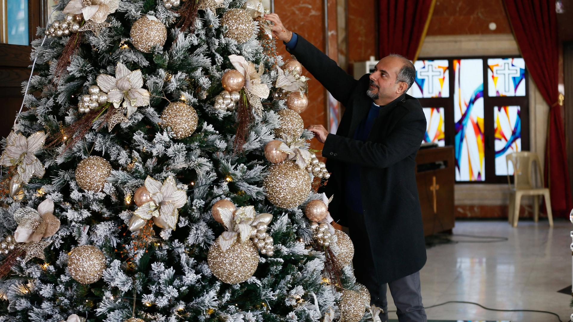 Ein Mann greift an einen festlich geschmückten Weihnachtsbaum, im Hintergrund ist das bunte Fenster einer Kirche zu erkennen.