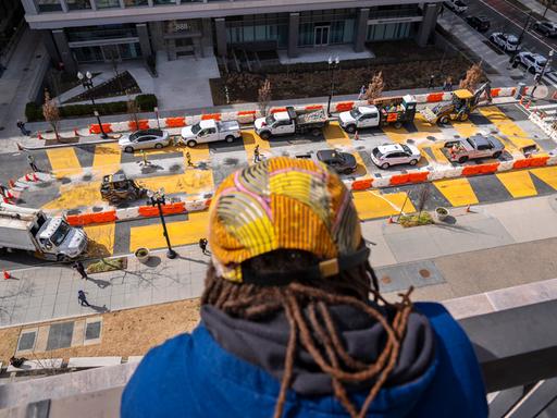 Eine Person schaut vom Dach aus auf eine Straße, in der in großen gelben Buchstaben noch  "Black Lives Matter" steht. Erste Absperrungen und Fahrzeuge bereiten dort die Zerstörung des Bildes vor.