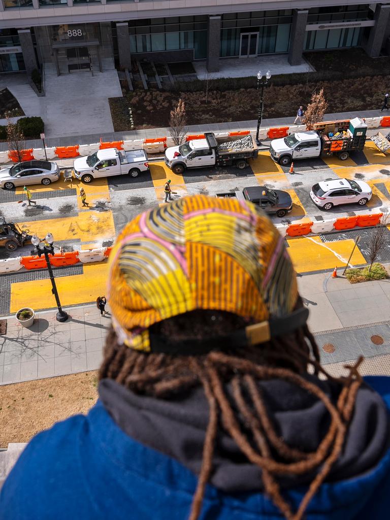 Eine Person schaut vom Dach aus auf eine Straße, in der in großen gelben Buchstaben noch  "Black Lives Matter" steht. Erste Absperrungen und Fahrzeuge bereiten dort die Zerstörung des Bildes vor.