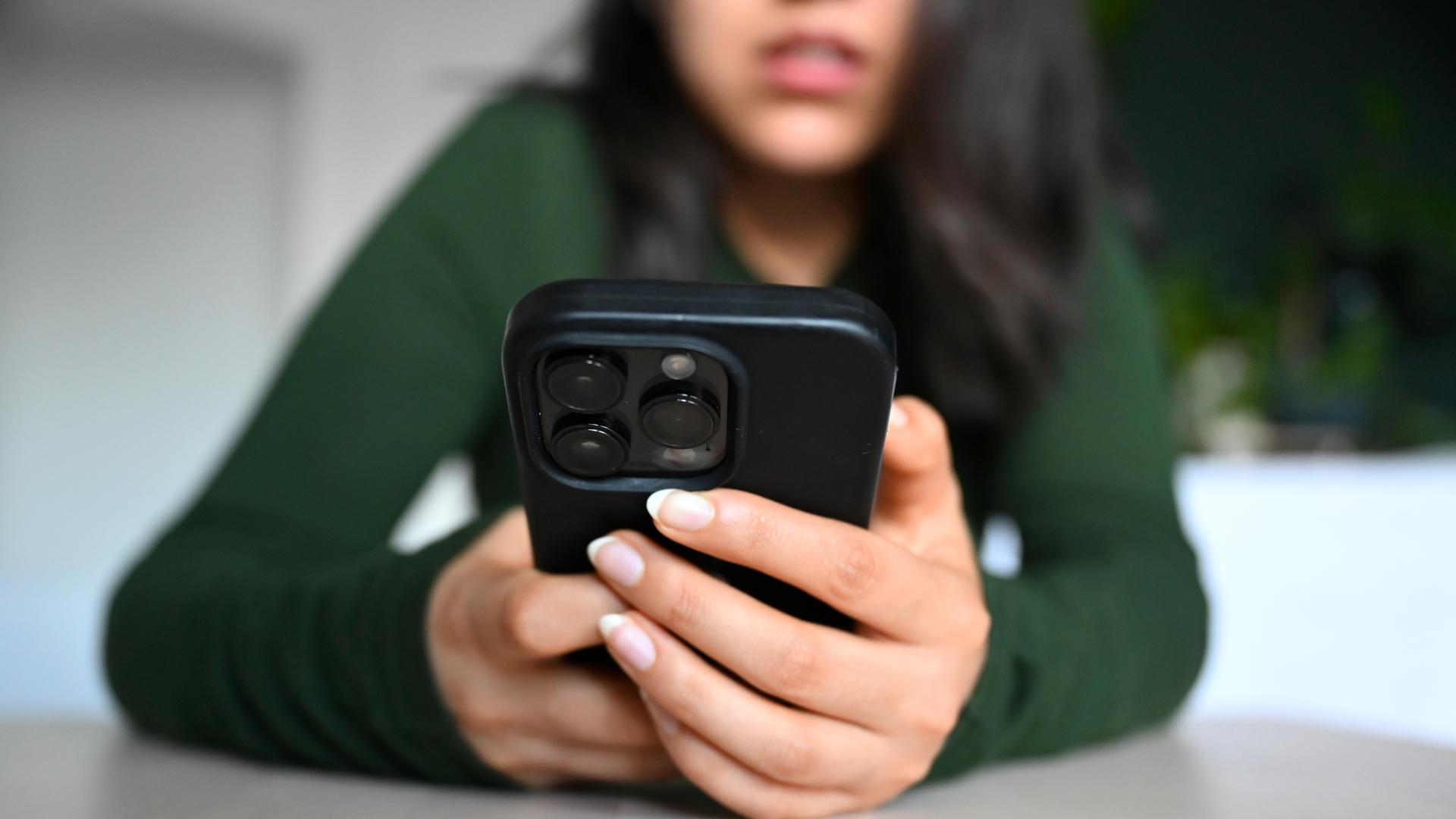 Eine junge Frau sitzt mit ihrem Handy an einem Tisch und blickt auf das Display.