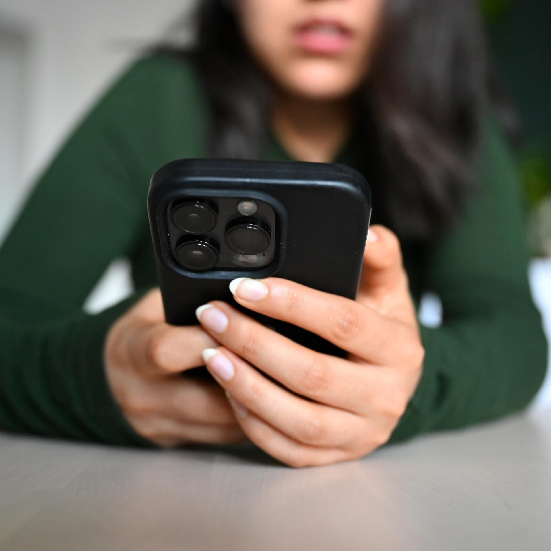 Eine junge Frau sitzt mit ihrem Handy an einem Tisch und blickt auf das Display.