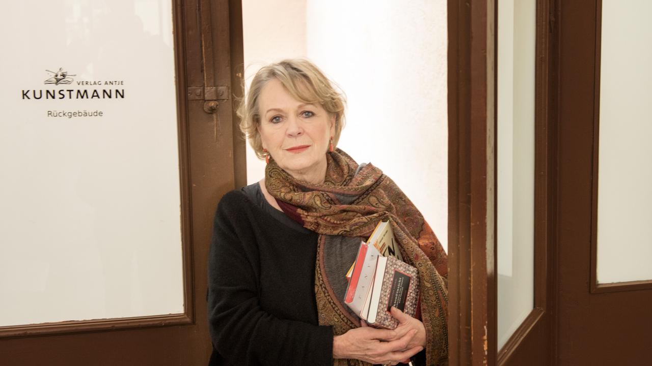 Antje Kunstmann steht vor einer Tür mit der Aufschrift "Verlag Antje Kunstmann" und hält einen Stapel Bücher unter dem Arm.