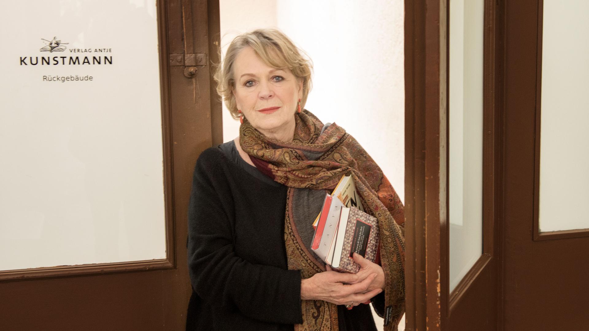 Antje Kunstmann steht vor einer Tür mit der Aufschrift "Verlag Antje Kunstmann" und hält einen Stapel Bücher unter dem Arm.