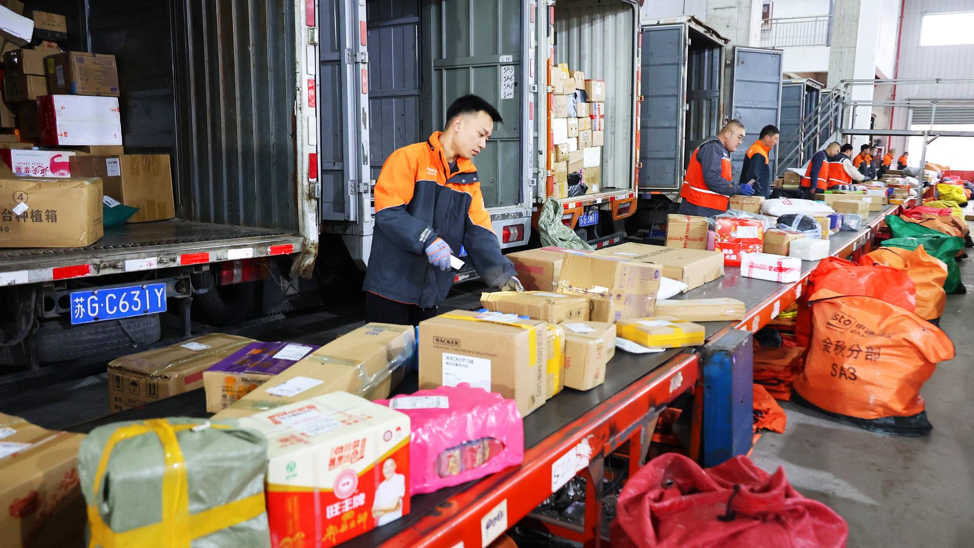 Mitarbeiter sortieren Expresspakete auf automatisierten Sortierlinien in einem Verteilzentrum in China, dahinter sind geöffnete LKW für den Transport.