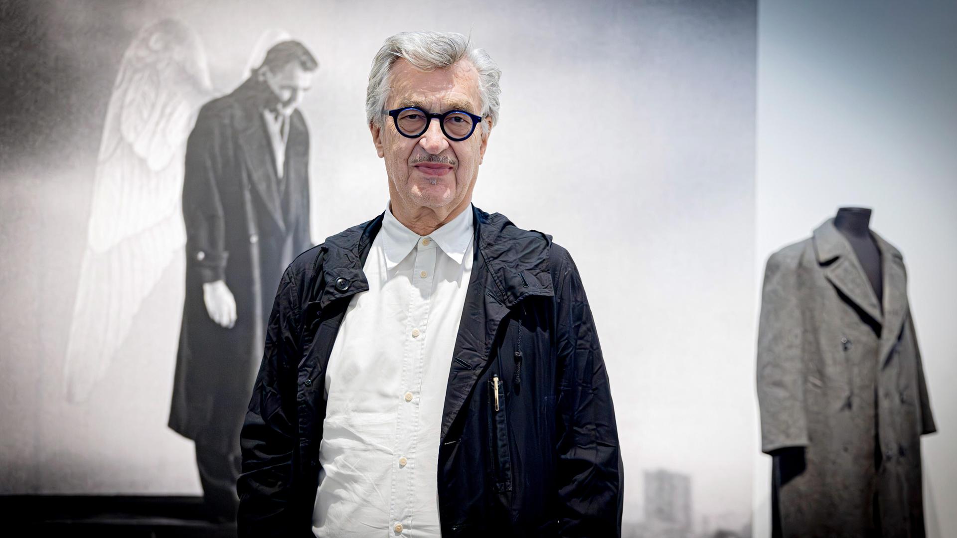 Wim Wenders steht vor einer Leinwand auf der ein Filmstill aus "Der Himmel über Berlin" zu sehen ist. Es zeigt Bruno Ganz, der mit Engelsflügeln am Rücken seines Mantels melancholisch auf Berlin blickt