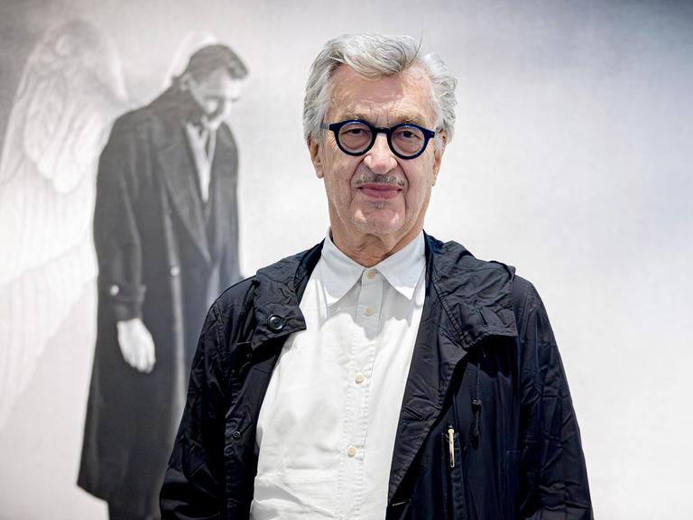 Wim Wenders steht vor einer Leinwand auf der ein Filmstill aus "Der Himmel über Berlin" zu sehen ist. Es zeigt Bruno Ganz, der mit Engelsflügeln am Rücken seines Mantels melancholisch auf Berlin blickt