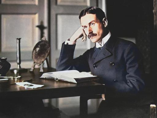 Thomas Mann sitzt an einem Tisch, den Kopf dabei in Hand gestützt. Nachkolorierte Fotografie.