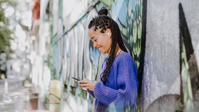 Eine junge Frau mit Kopfhörern und Smartphone steht lächelnd an eine Wand mit Graffitis gelehnt
